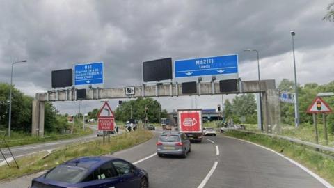 An approach to Simister Island (junction 18) on the M60 motorway. There are only a few cars and a van on the road which splits in two directions with signage on a concrete goalpost type structure across the entire carriageway. Left is for the M60 and M62 west to Liverpool. Right is for the M62 east to Leeds. There are traffic lights and a large roundabout in the distance.