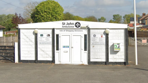 A single-storey white structure with the words St John Ambulance Isle of Sheppey Divisions in black writing above the front door, in the centre of the structure