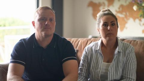 Jack and Lindsey Kitching are sitting down on a brown couch in their home. They both have serious expressions on their faces and are starring off camera.