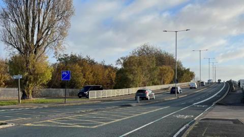Hessle Road flyover with cars driving over it. It has on-road cycle lanes and street lights along the central reservation. There are trees to the left and a blue sign for a cycle lane.