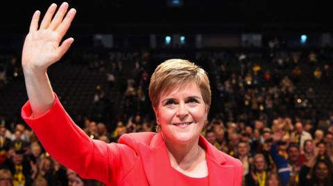 A woman, photographed from the chest up, looking up and waving with her right hand. She has short brown hair, combed to the side, and is wearing a red top and matching red jacket. A large crowd of people are visible in the background in the image, which was taken in an auditorium. 