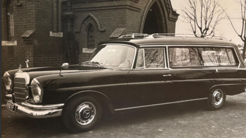 A black and white photo of a Rolls-Royce hearse standing outside a large brick building