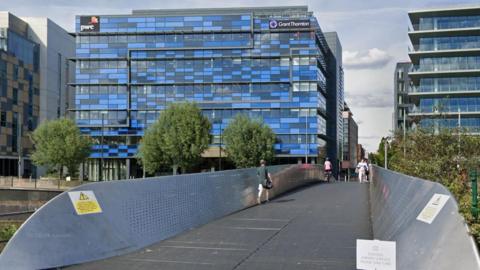 A screenshot from Google street view showing the metal bridge with thousands of small holes on either walled side. There are pedestrians and one cyclist crossing the bridge. In the background there is a large office building with lots of windows and blue panelling. 