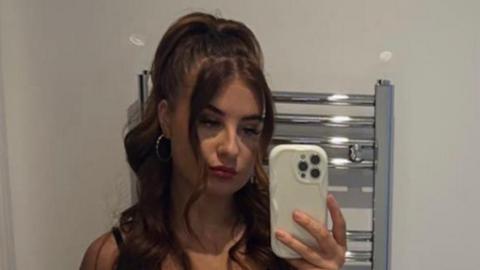 Paige Roberts taking a selfie in a mirror, she has long wavy dark brown hair and large hooped earrings. Her iPhone is visible and behind her is a vertical, wall-mounted bathroom radiator 