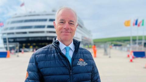 Bob Sanguinetti, a man standing at a harbour, wearing a Port of Aberdeen-branded jacket, with a cruise ship in the background.