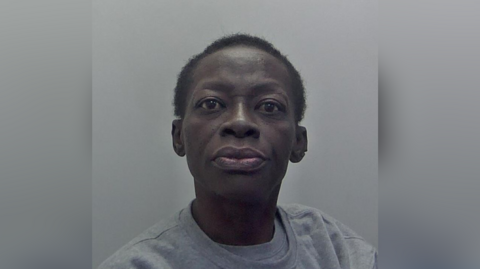 A police mugshot of a woman in a grey jumper with shaved hair. 