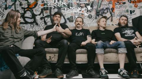 Pigs x7 which is made up of five men, all sitting on a sofa in front of a graffitied wall. They are all wearing dark colours and the two on the left are holding their thumbs together like they're in the middle of a thumb war. The man on the far left has long, brown hair and is looking to the side. Next to him, the man has a grey beard and is wearing a black cap. The man in the middle has dark hair which looks to be tied up and a dark moustache. He is wearing a black Korn band t-shirt. The man middle right has light hair and is wearing a grey cap and is looking to the right with his hands in his lap. He is wearing turned up jeans that are exposing the bottoms of his legs. The band member on the far right has chin-length dark hair and a greying beard and is looking into the camera with his hands clasped on his lap.