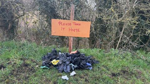 Dozens of black bags dumped under a homemade sign reading 'please take it home'. They are on a grassy bank near a hedgerow.