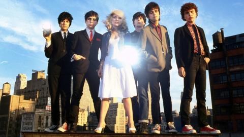 Members of Blondie stand together on a rooftop in New York City in 1978, with Debbie Harry in the centre and a camera flash reflecting off her white dress.