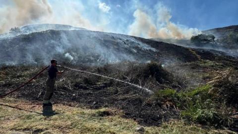 A firefighter spraying water on moorland with smoke coming out of the ground