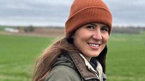 A woman in a beanie hat and a green coat stands in a field. She is smiling at the camera.