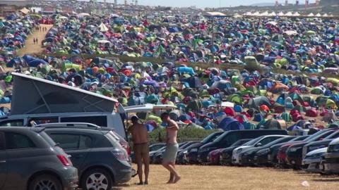 A photo of the festival with thousands of tents over the hills in Newquay. Two men are shirtless and opening the back of a campervan with hundreds of cars parked below the tents.