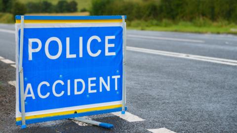 A blue and white 'police accident' sign at the side of a single carriageway, lined with greenery.