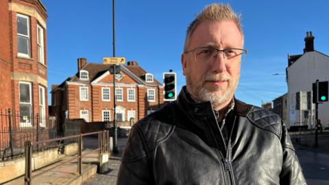 Ian Milligan looks directly at the camera. He is wearing a black leather jacket, has glasses on and has a white beard. He is standing on a pavement outside a Georgian red-brick building, with 11 sash windows to its front and a blue door. The sky is clear blue.