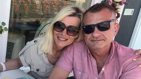 A man and woman wearing dark glasses sit at a cafe table. The blonde woman has her arm around the man who is wearing a pink short-sleeved shirt. 