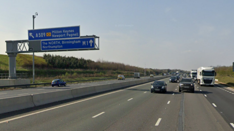 A Google Maps street view image of the M1 between junctions 14 and 15. Vehicles are travelling on the road in both directions and a blue sign directing drivers towards Milton Keynes and the North can be seen on the left. 