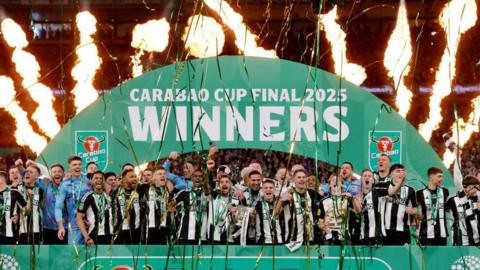 Newcastle United's Bruno Guimaraes and Kieran Trippier hold the trophy after winning the Carabao Cup. The squad, with their medals, surround them on a green stage on the Wembley pitch. An arch above them carries the message Carabao Cup Final 2025 Winners. Behind them are jets of flame while streamers fill the air in front.