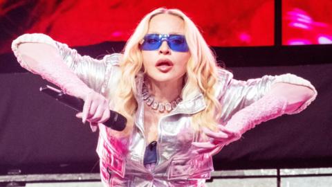 Madonna on stage in New York. She's holding her arms to the side, revealing pink lace gloves beneath a silver jacket. Her eyes are hidden behind jet blue sunglasses, but she appears to be staring directly at the camera.
