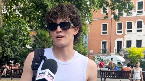 A young man wearing sunglasses and a white vest speaking into a BBC microphone in a park