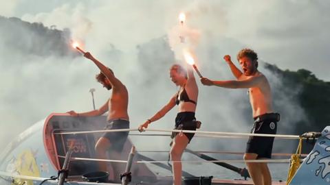 Three rowers stand on top of boat in swimwear holding flaming flares aloft in celebration