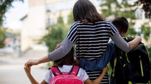 Woman walking with two children pictured from the back. The girl is carrying a pink shoulder bag and the boy a black and green back and the woman has her arms around the childrene 