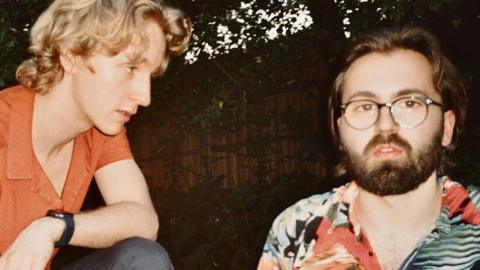 Two men, one is blond with an orange polo on, while the other has brown hair with a beard and glasses.