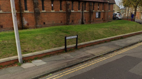 A street view image of a road with double yellow line and a pavement. A street name sign is visible with a grass bank behind it and a red brick building.