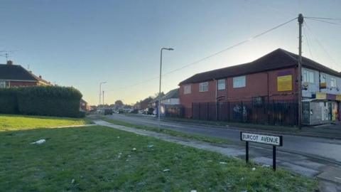 A sign for Burcot Avenue is in the foreground, with the road going off into the distance, with a grassy area on the left and red-brick properties on the right hand side.