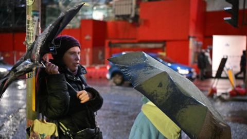 Two people talking under umbrellas. The one on the left is a woman wearing a black coat, maroon woolly hat and she is carrying a black umbrella that is being pulled inside out by the wind. She has a black handbag over her shoulder. The second person has their back to the camera and is wearing a green and yellow jacket. A black umbrella is obscuring their face. There are cars driving past red boards behind them.