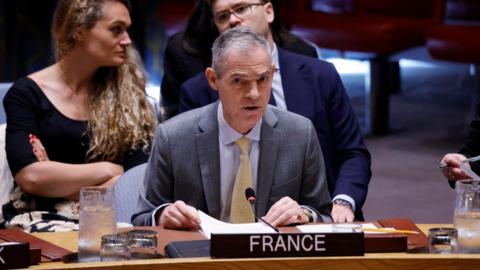 France's UN Ambassador Jerome Bonnafont speaking at the UN Security Council on the table with a "France" placard while two people sit behind him