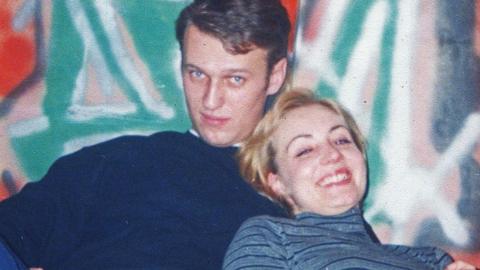 Alexei Navalny and his wife Yulia in their youth, the photo is cropped tight showing their head and shoulders as they embrace in front of a graffiti'ed wall