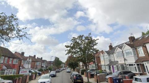 Jubilee Road in Perivale, London, with terraced houses on either side of the street and a tree on the right hand side. Cars also line the streets and the sky is blue with white clouds.