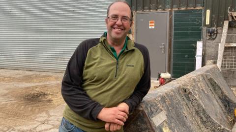 James Winslade is leaning against a tractor in front of a farm building. He is wearing a green and black top and glasses. He is smiling.