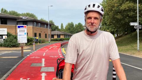 Jim wearing a cream T-shirt and a white helmet on his bicycle - along the red cycle path on Thorpe Wood 