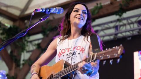 Amy Macdonald performing onstage, wearing a white T-shirt and playing an acoustic guitar.
