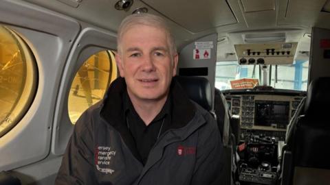 Ryan McNay, pictured onboard the emergency transfer plane. The flightdeck is visible behind him. He has grey hair, a black jacket and black shirt. The plane windows are visible over his shoulders.