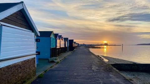 A row of beach huts in Hamworthy next to the promenade and harbour under a cloudy sky with a low sun reflecting on the water