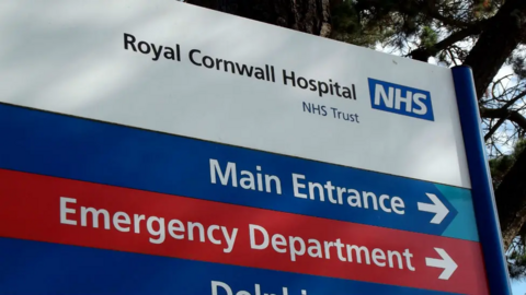 A red, white and blue sign in the grounds of the Royal Cornwall Hospital showing directions to the main entrance and the emergency department