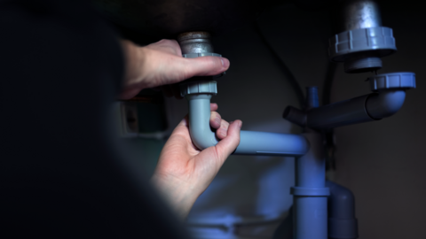 A stock image showing a pair of hands holding on to some some pipes, seemingly under a sink or in a cupboard.