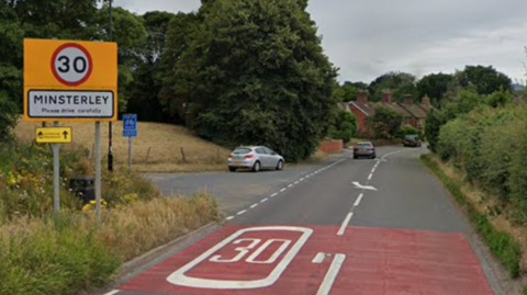 A road leads into a village, and to the left is a yellow sign with 30mph on it, and the words Minsterley, please drive carefully. Houses can be seen on a bend in the distance