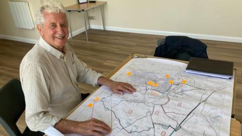 A man is smiling at the camera. He is sitting in front of a map with lots of notes and orange stickers.