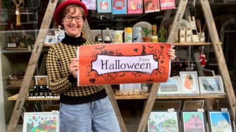 Nicola Bartlett outside her Eclectic Gift Shop holding an orange banner which advertises the halloween trail.