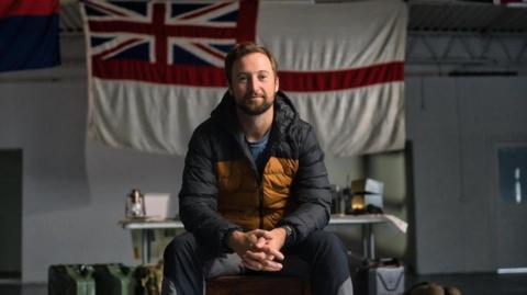 Fred Stokes poses for a photo sitting on a chair, his hands clasped, wearing a black and yellow padded jacket. Behind him is an England flag with the Union Jack in the top left corner. A table with equipment can be seen in the background. 
