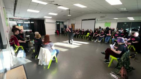 Inside a rehearsal room, a number of performers sit down on green chairs in front of some mirrors. They are all wearing black T-shirts that says "All Stars Theatre Group". One man is standing the middle of the room, holding a microphone and singing. Several people are also sat at a desk.