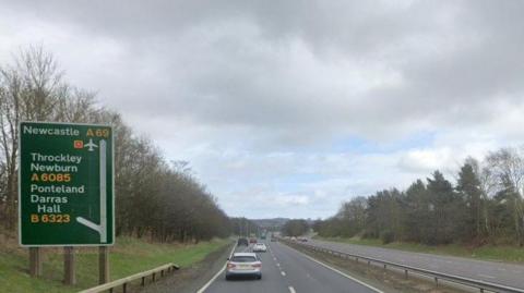 The A69. There is a green sign on the left, giving directions for Newcastle, Throckley, Newburn, Ponteland and Darras Hall. A grey and a white car are travelling in the left lane, with more vehicles further ahead. 