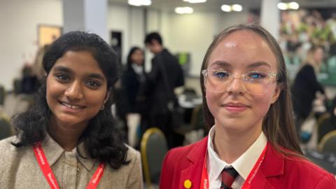 Two girls standing in a conference centre. the girl on the left is dressed in a beige blazer, the girl on the right is wearing a red blazer. They are smiling.