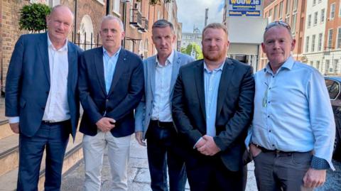 Five men stood on a street, most of whom are looking at the camera. They are all wearing shirts and most are wearing suit jackets. 