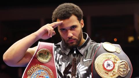 Moses Itauma seen from chest up looks into the camera as he tilts his head forward slightly and salutes with two fingers on his right hand raised to his forehead. He is wearing a black leather waistcoat with a white design on it and has a boxing belt, one red and one black, draped over each shoulder