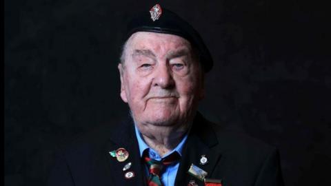 Richard Aldred sits with his hands clasped in front of him. He is wearing a dark blue jacket with blue shirt and green, red and gold tie. His jacket is decorated with medals and badges. The background behind him is black.