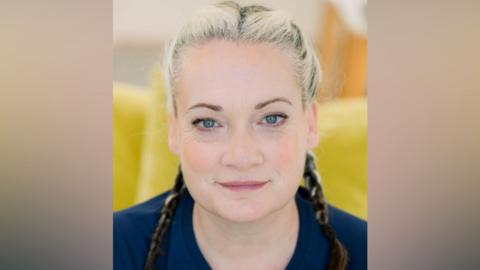 A woman smiles at the camera. She has blonde hair which has been French braided. She is wearing a dark blue T-shirt.